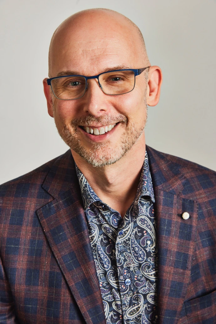 A headshot of Mike. His head is shaved. He has brown and grey beard. He is wearing blue glasses, a red and blue plaid blazer, and a blue and white paisley collared shirt.